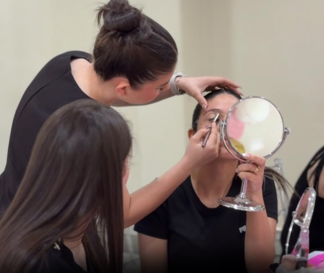 Una profesora de la academia guía a una alumna que se maquilla los ojos frente a un espejo, en plena clase